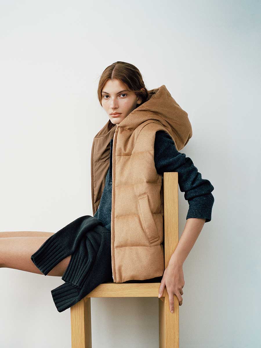 Das sitzende Model trägt eine wattierte Max Mara The Cube Weste in Kamel mit zum Look passender Kapuze in Dunkelblau.